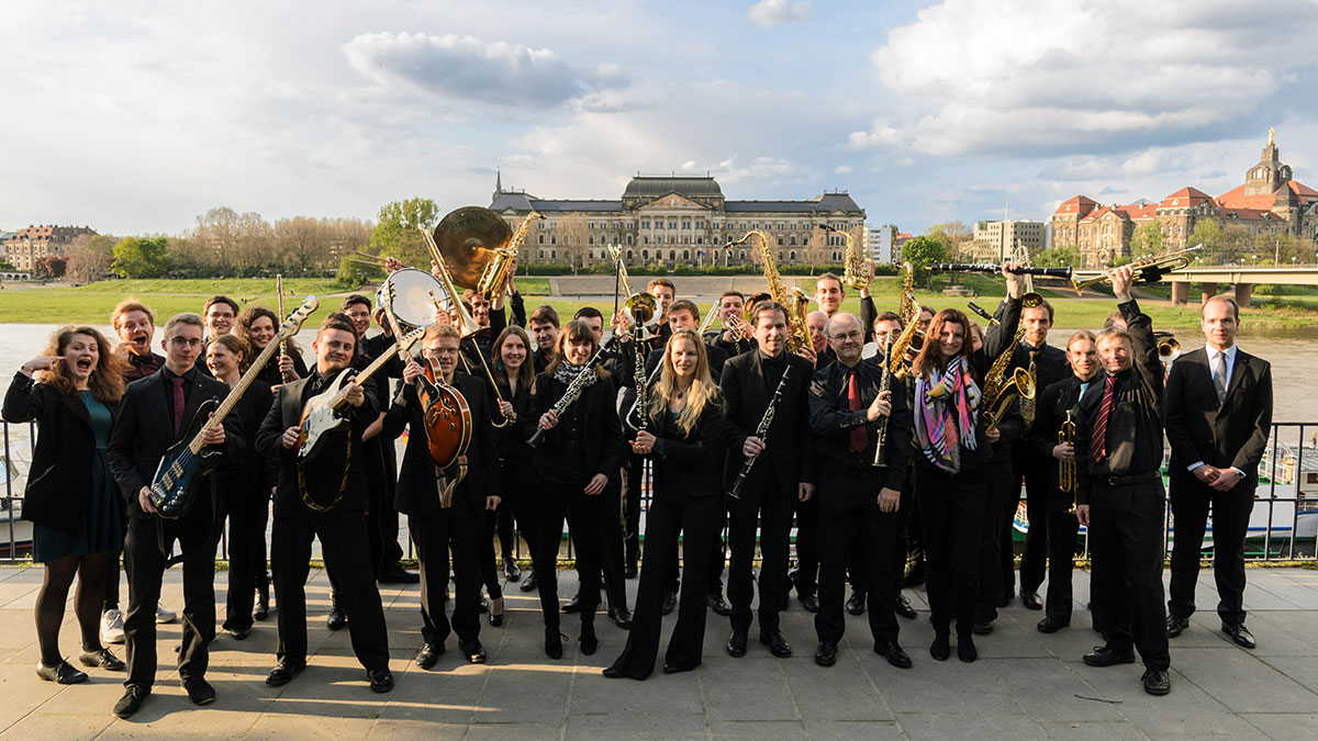 Eine Gruppe von mehr als 30 schwarz gekleideten Personen präsentieren ihre Instrumente. Im Hintergrund ist die Elbe und das Sächsisches Staatsministerium für Kultus zu sehen.