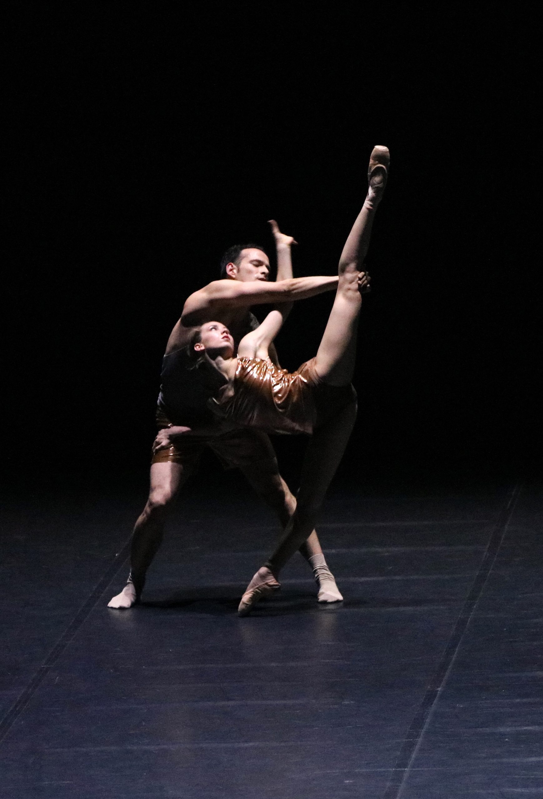 Zwei Balletttänzer tanzen ein Duett. Sie tragen goldene Kostüme und der Hintergrund ist dunkel.