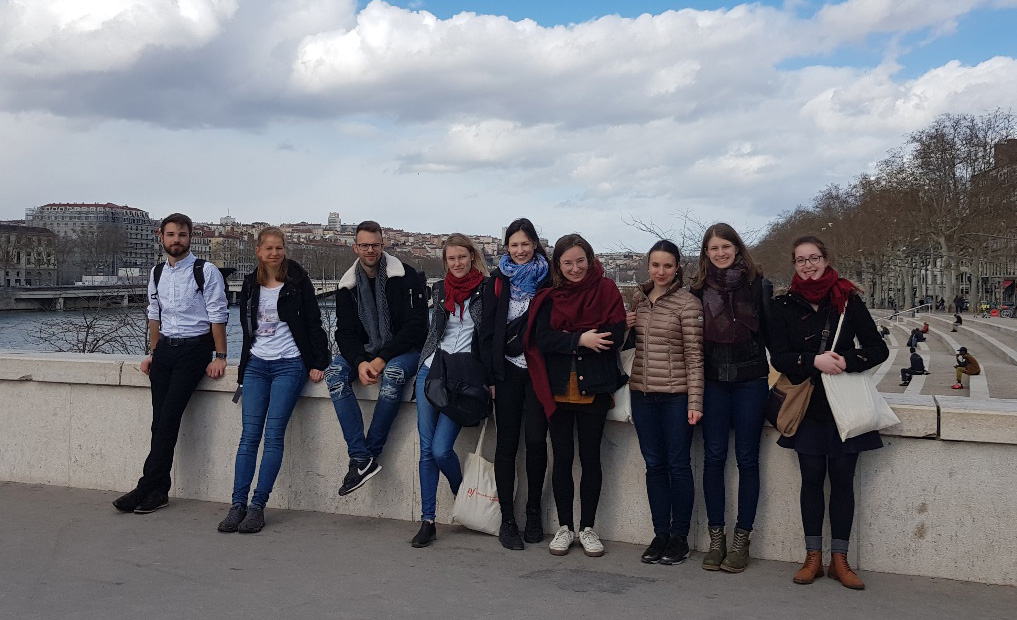 Eine Gruppe von 9 Studentinnen und Studenten steht auf einer Brücke über einen Fluss. Im Hintergrund sieht man die Stadt Lyon.