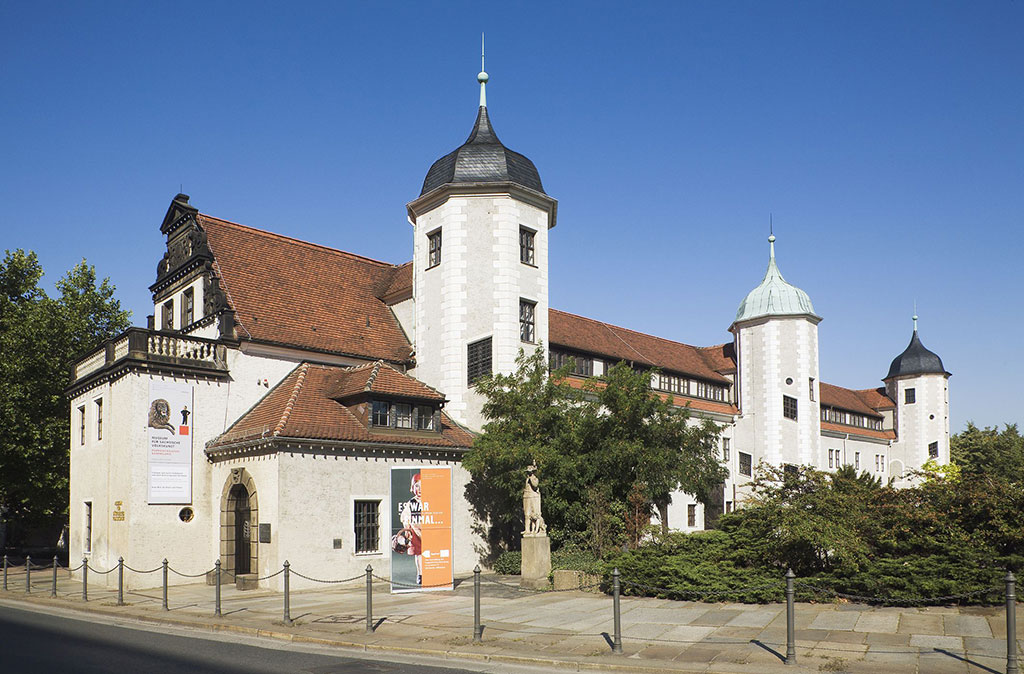Dieses Foto zeigt den Jägerhof Dresden, ein altes Gebäude mit Spitzdach und drei Türmchen an der langen Seite rechts daneben gibt es Sträucher und Bäume. Es beherbergt das Museum für Sächsische Volkskunst Dresden.