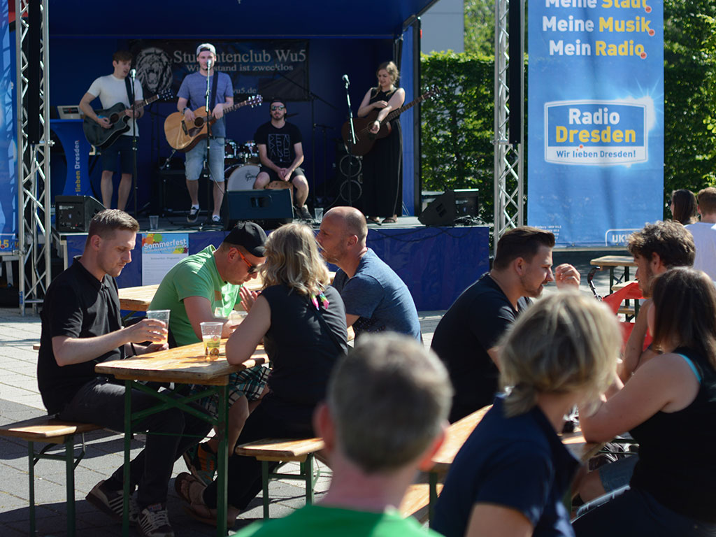 Mehrere Studenten sitzen mit kühlen Getränken an Biertischgarnituren, während auf der Bühne von Radio Dresden eine Band spielt. Aufnahme vom Sommerfest Wundtstraße aus dem Jahr 2017.