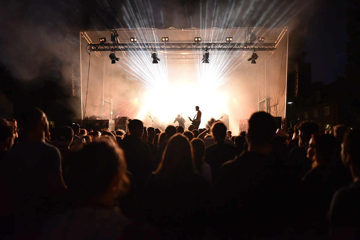 Foto: Eine Band spielt auf einer beleuchteten Bühne am Abend mit einer großen Anzahl von Zuschauern.