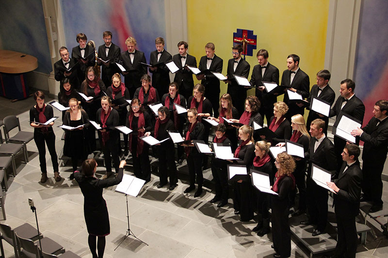 Das Bild ist eine Fotografie des Kammerchors der Tu Dresden. In der linken unteren Ecke steht die Dirigenten und ihr gegenüber halbkreisförmig angeordnet die Sängerinnen und Sänger des Chores mit Notenbüchern in den Händen.