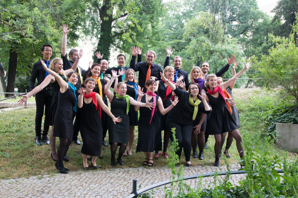 Das Foto des Chors Vokalwerk zeigt 12 Frauen und 7 Männer in schwarzer Kleidung mit bunten Krawatten und Schals, die in die laut lachen und die Hände heben.