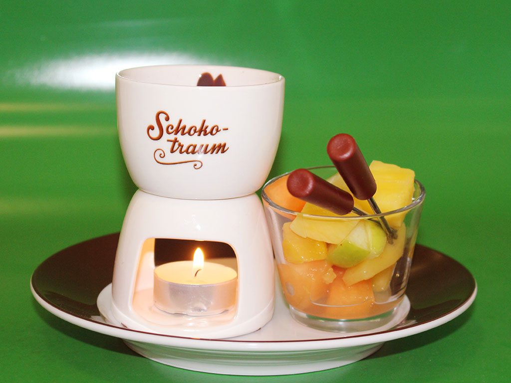 Abgebildet ist ein kleines Schokoladen-Fondue, daneben ein Glas mit Früchten und einem kleinen Spieß.