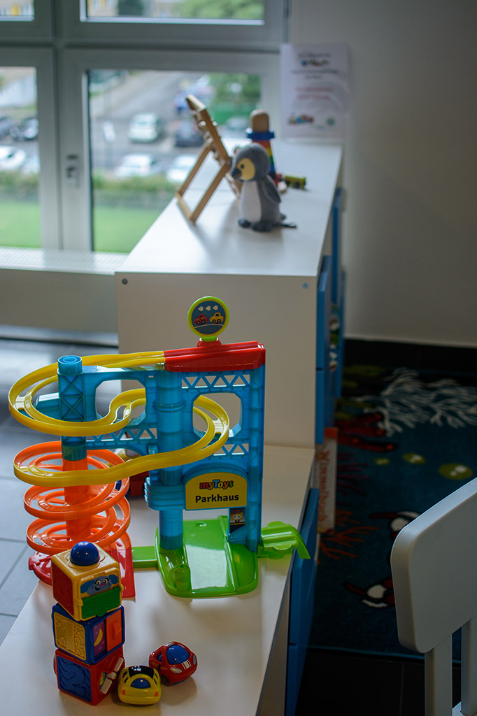 Das Bild zeigt ein Spielzeug-Parkhaus. Von oben führt eine spiralförmige Strecke nach unten, auf der Spielzeug-Autos fahren können. Davor stehen drei übereinandergestapelte Würfel und zwei Spielzeug-Autos. Im Hintergrund sieht man eine Speilzeugteppich, der zur Spielecke gehört.