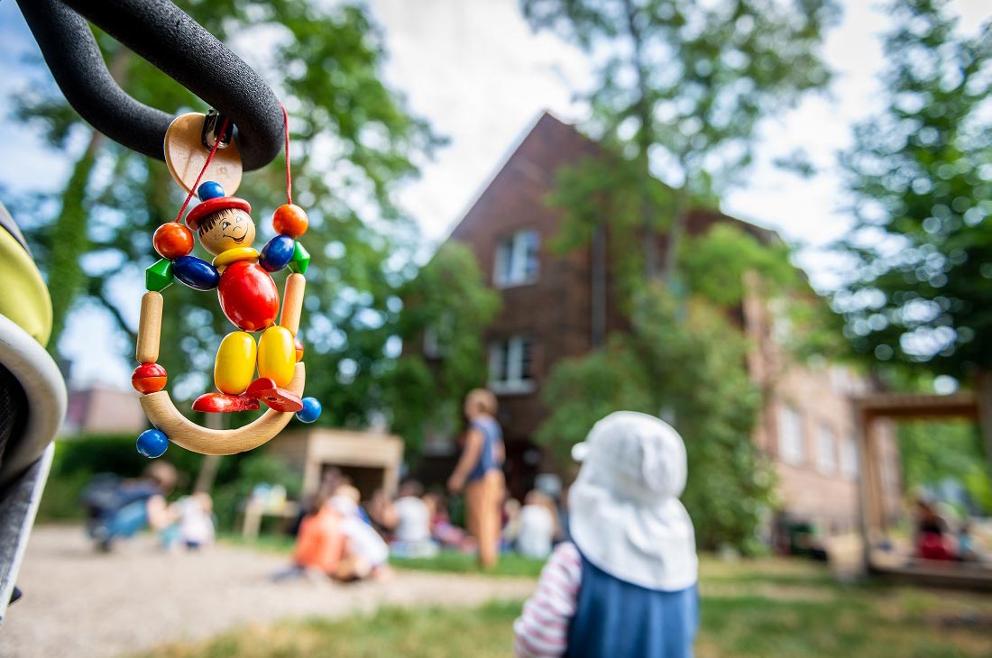Foto: Im Vordergrund ist ein Holzspielzeug und im Hintergrund eine Kindergartengruppe auf einem Spielplatz.