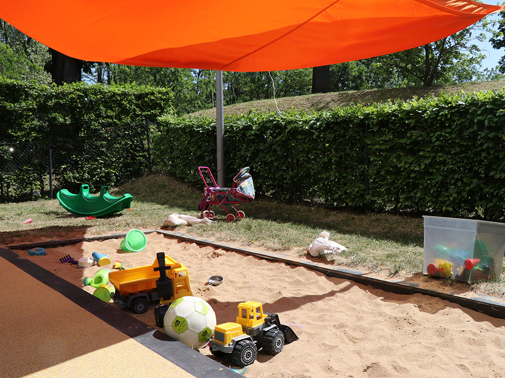 Im Sandkasten des Campusnests findet sich Sandspielzeug, ein Ball und mehrere Bagger sowie ein Puppenwagen, der neben dem Sandkasten steht.