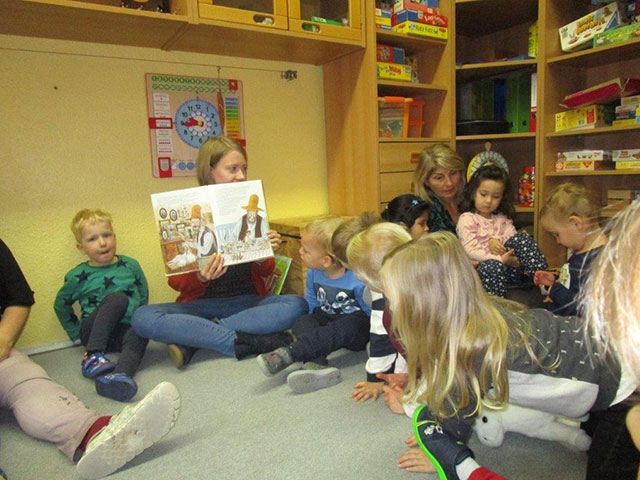 Mehrere Kinder sitzen auf dem Fußboden und hören einer Frau, die aus einem Buch vorliest, gespannt zu.