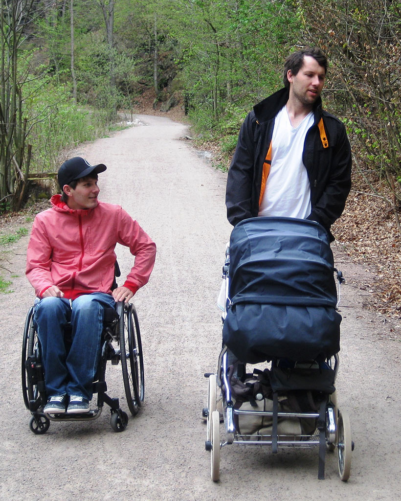 Passend zum Artikel mit dem Programm der diesjährigen Diversity Tage an der TU Dresden zeigt dieses Foto zwei junge Männer auf einem Waldweg nebeneinander. Der linke Mann trägt ein Base Cap und sitzt im Rollstuhl. Der rechte Mann schiebt einen Kinderwagen.