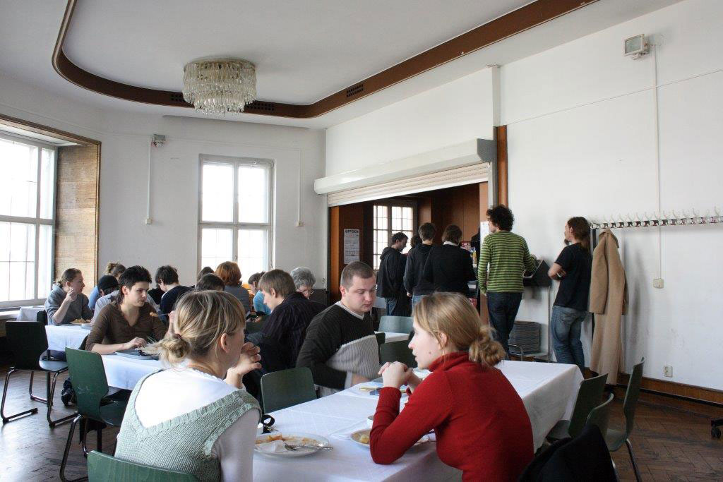 Foto eines Speisesaals: mehrere Personen sitzen an Tischen und essen. Im Hintergrund stehen einige Personen an der Essensausgabe an.