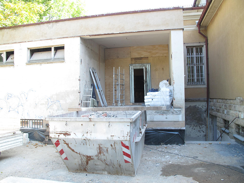 Foto des Hintereingang mit unverputzen Wänden und einem Container für Bauschutt