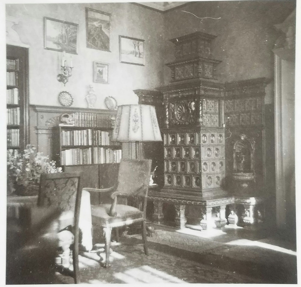 Altes schwarz-weiß Foto einer Zimmerecke mit einem Kachelofen, mehreren Bücherregalen und einer Lampe.