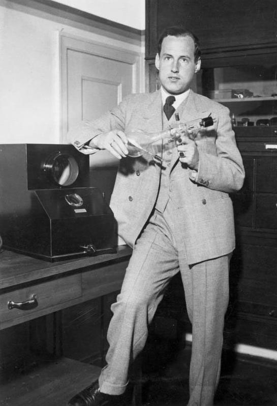 schwarz-weiß Foto von Manfred von Ardenne der in einem Raum steht und ein elektrisches Geräte in den Händen hält.