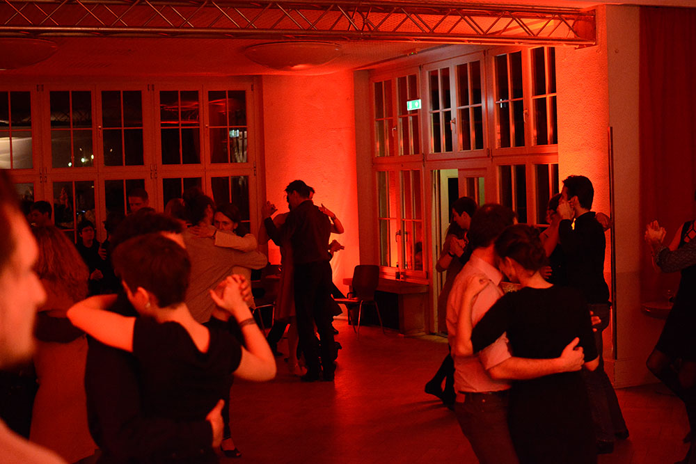 Foto eines rot beleuchteten Saal. Mehrere Paare tanzen Tango.